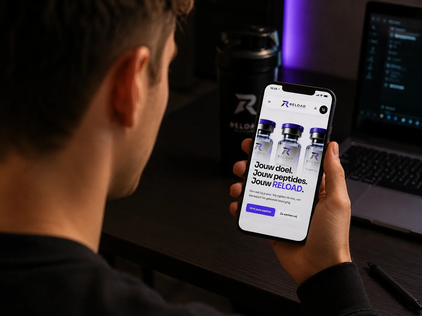 Iemand bekijkt de RELOAD-website op een telefoon aan een bureau, met een shaker en een laptop op de achtergrond